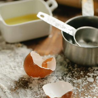 Close-up of broken eggshells, measuring cups, and a bowl on a floured kitchen countertop.