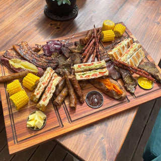 A beautifully arranged braai platter featuring meats, grilled corn, and toasted sandwiches on a wooden board.