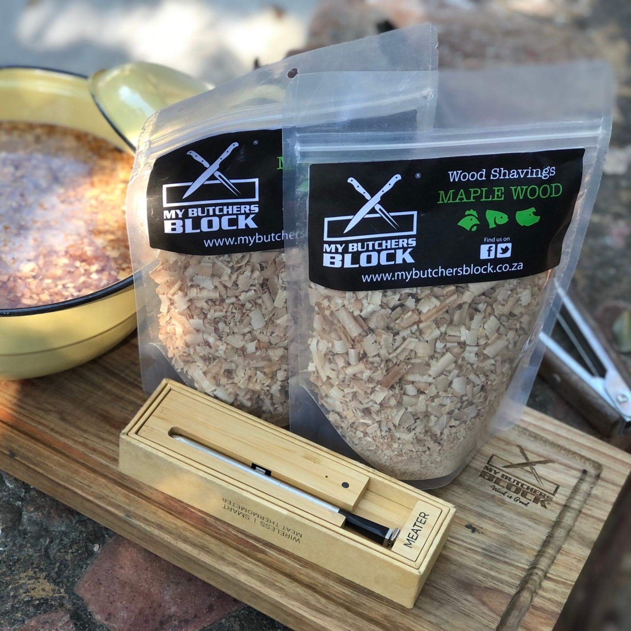Premium maple wood shavings for smoking food, displayed in clear bags on a wooden board with a smoking thermometer.