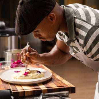Chef garnishing a creamy dish with edible flowers on a wooden cutting board.