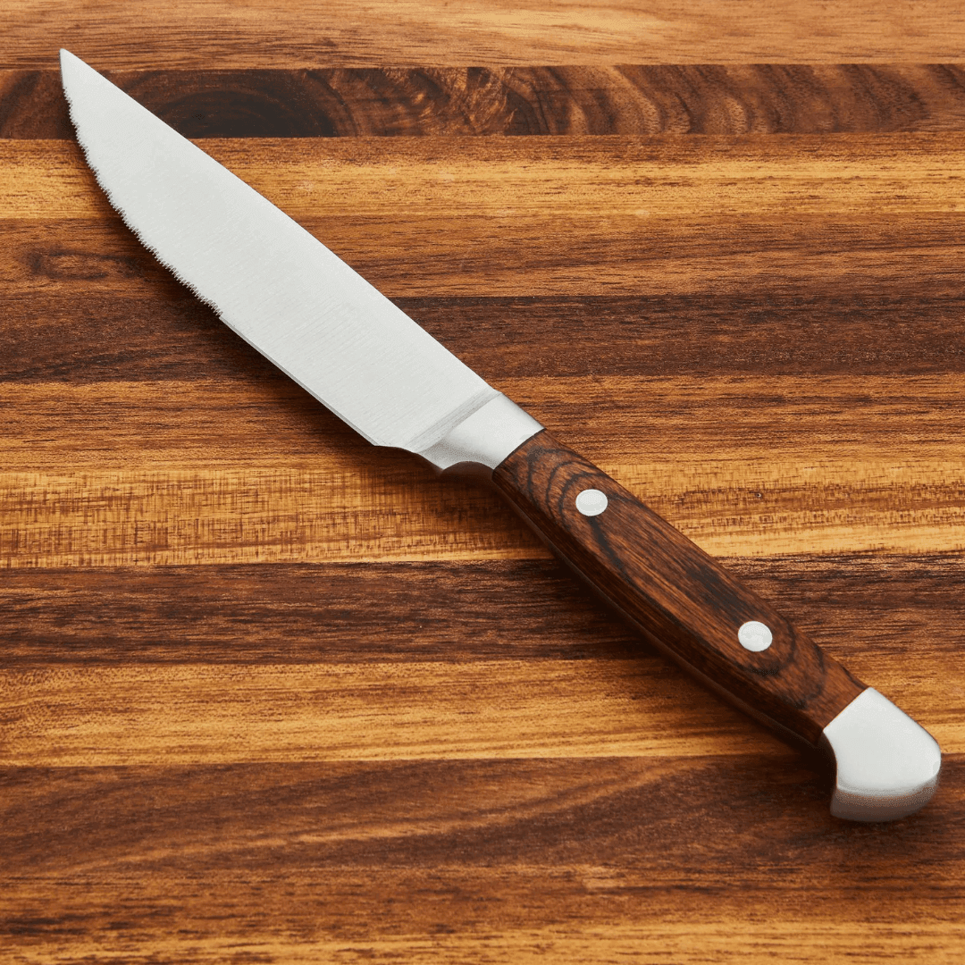 Close-up of a steak knife with a serrated stainless steel blade and a wooden handle on a wooden surface.