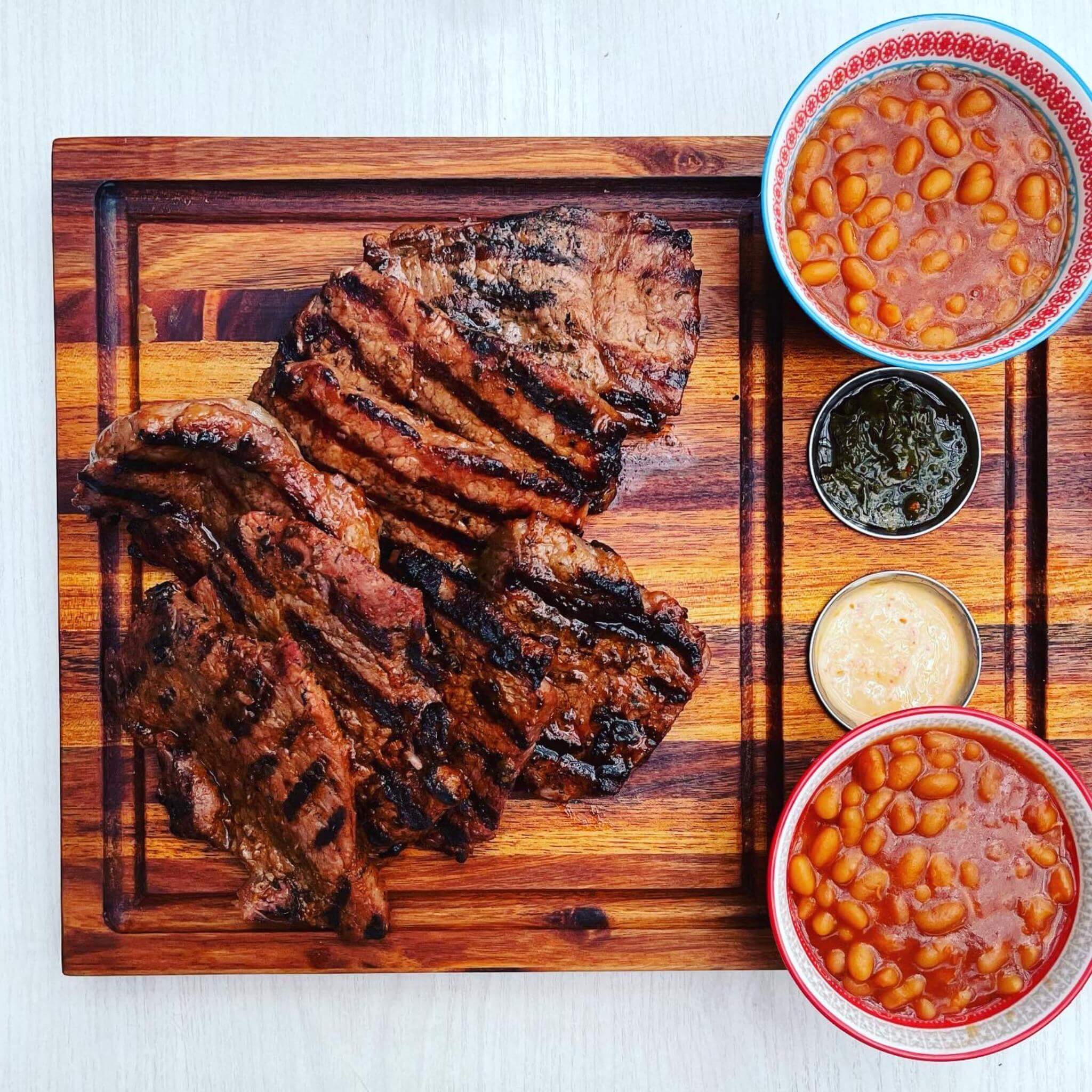 Grilled steaks on a wooden meat board with bowls of beans and sauces for an outdoor braai experience.