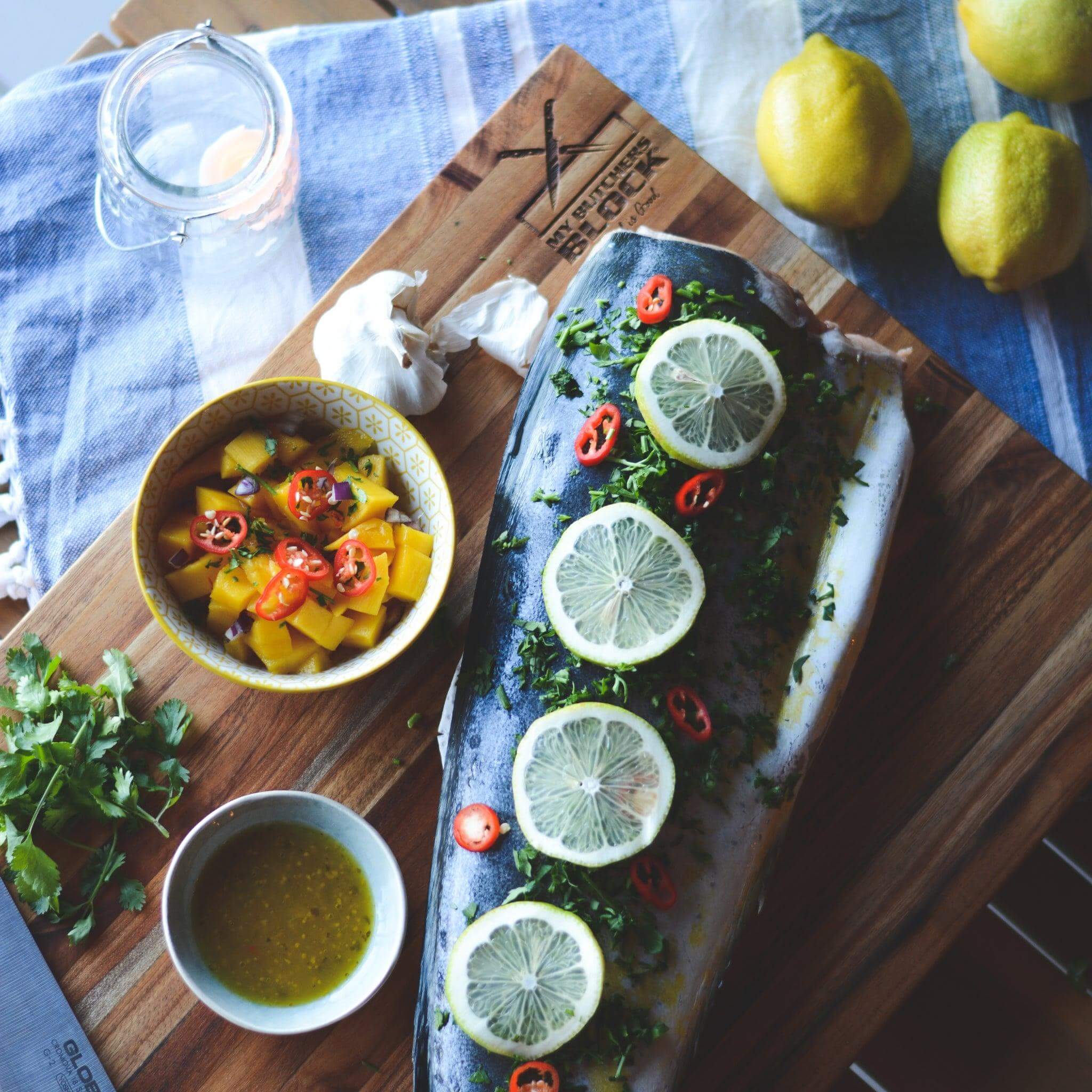 Large wooden chopping block with seasoned fish, lemon, chili, and sauces, perfect for South African cuisine.