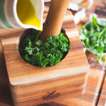 Fresh parsley being mashed in a wooden mortar with olive oil pouring from a jug, ideal for culinary use.