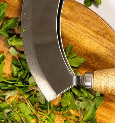 Close-up of a stainless steel herb knife on a wooden cutting board with fresh chopped herbs.