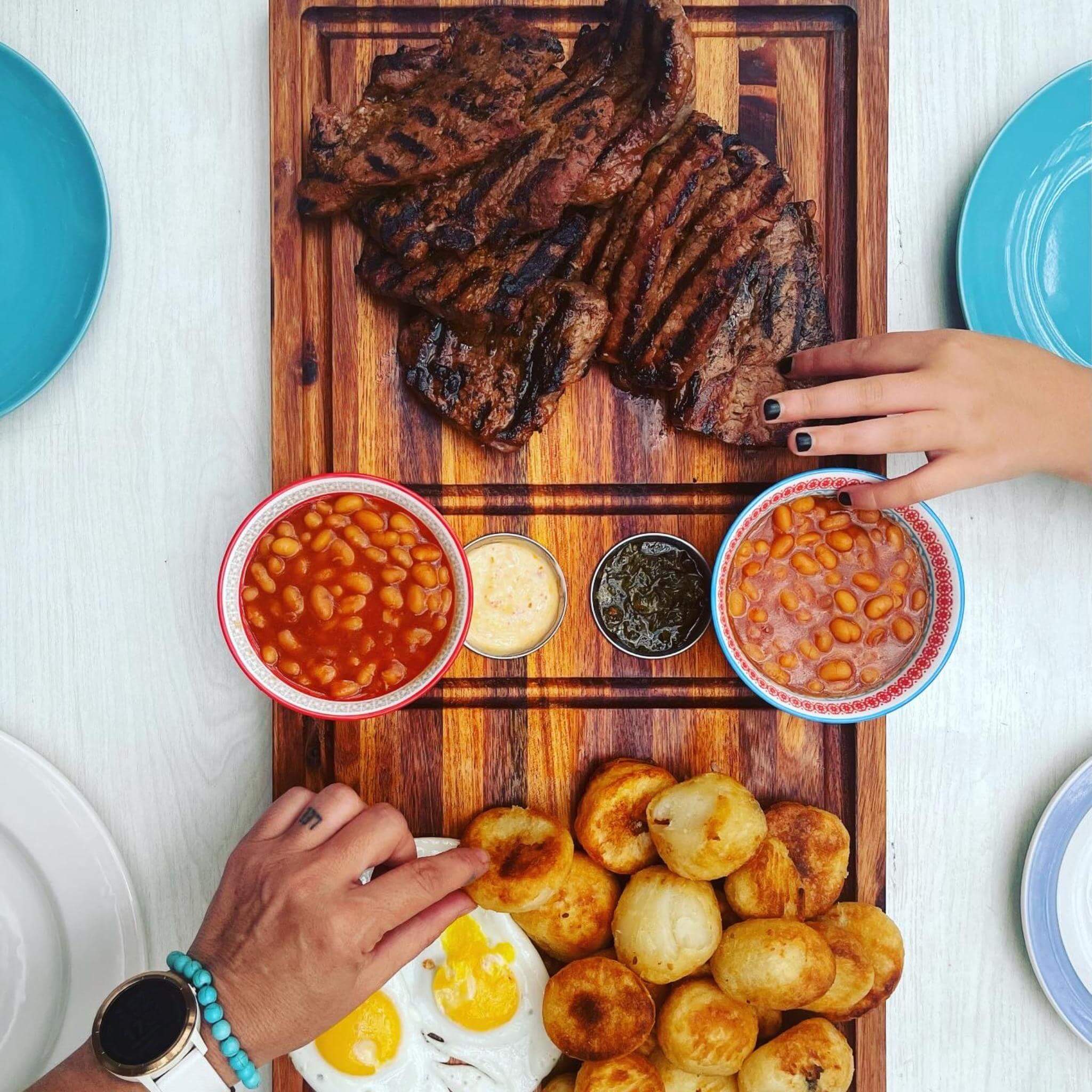 Delicious grilled steak on a wooden platter with sides, including beans and golden bread rolls, ready for serving.