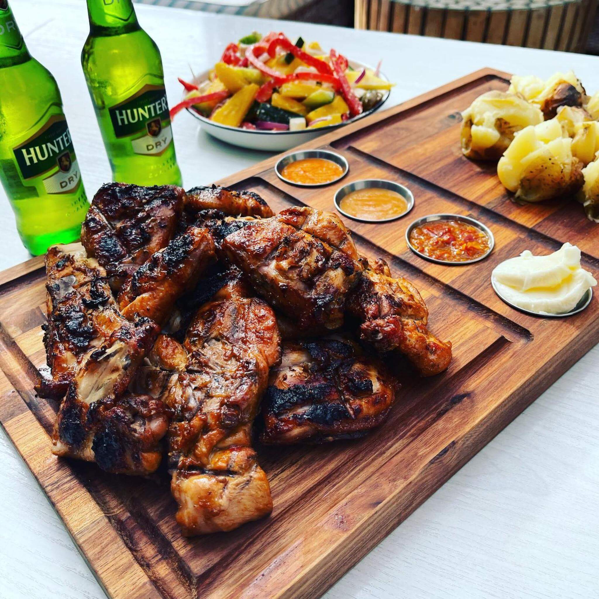 Grilled meat platter with sauces, fresh salad, and drinks on a wooden board for an outdoor braai experience.