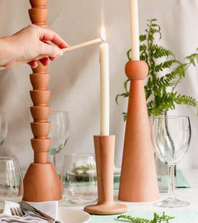 Hand lighting a taper candle in elegant wooden candle holders, surrounded by greenery and glassware on a dining table.