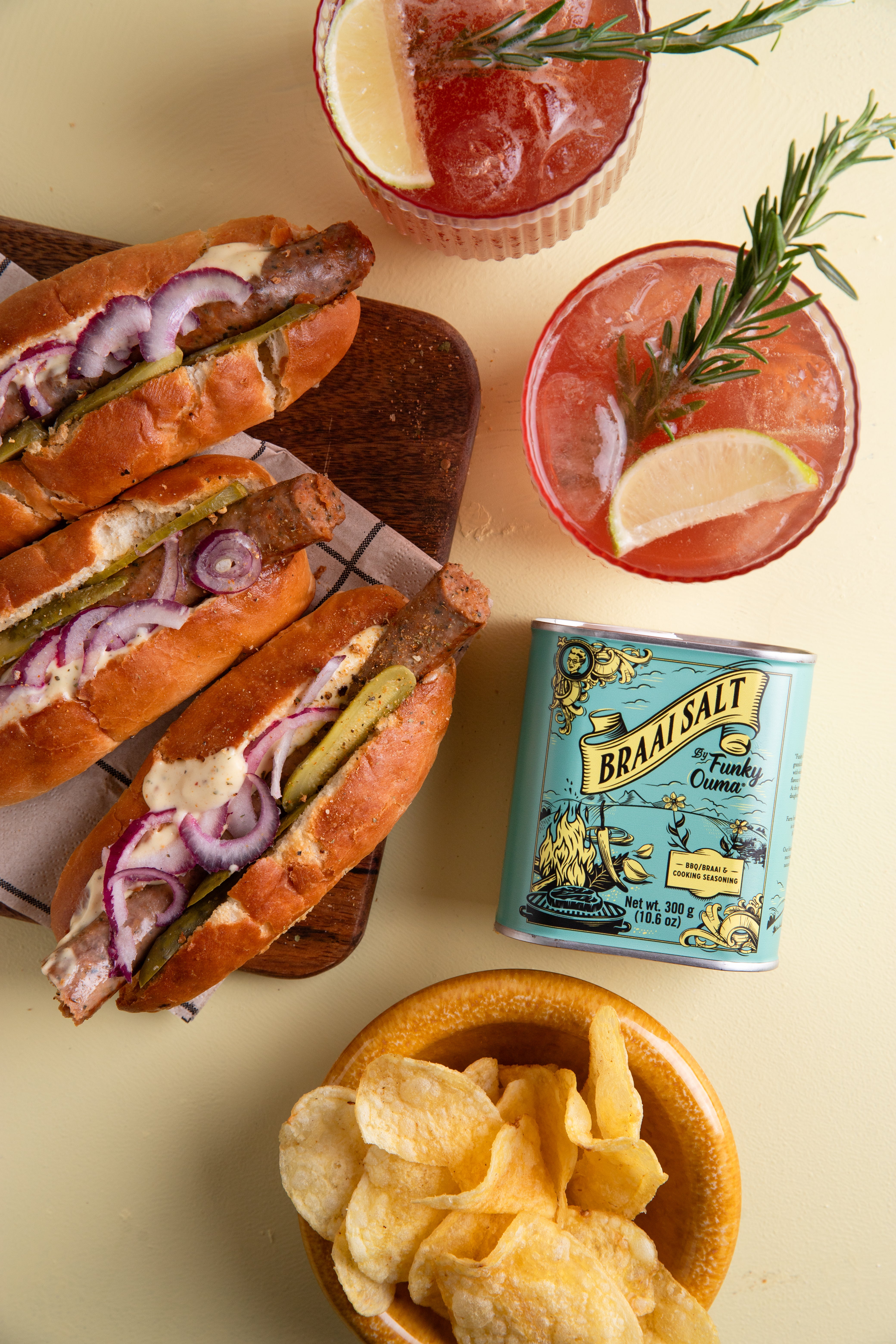 Savory braai hot dogs with onions, pickles, cocktails, and chips on a table.