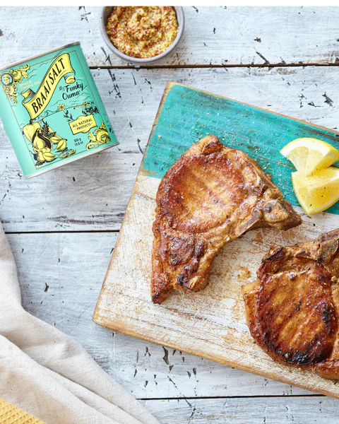 Grilled pork chops on a chopping block with Funky Ouma Braai Salt and lemon slices.