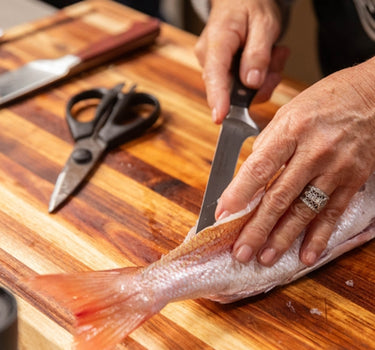 Close-up of hands preparing fresh fish on a wooden cutting board with kitchen knives and scissors.