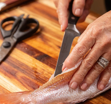 A person skillfully filleting a fish on a wooden cutting board with kitchen knives and scissors nearby.