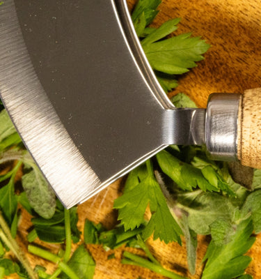 Herbs being chopped on a wooden cutting board with a stainless steel herb chopper.