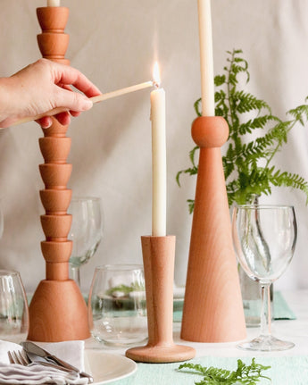 Hand lighting a white candle on elegant wooden candle holders, with fern leaves and glassware in the backdrop.