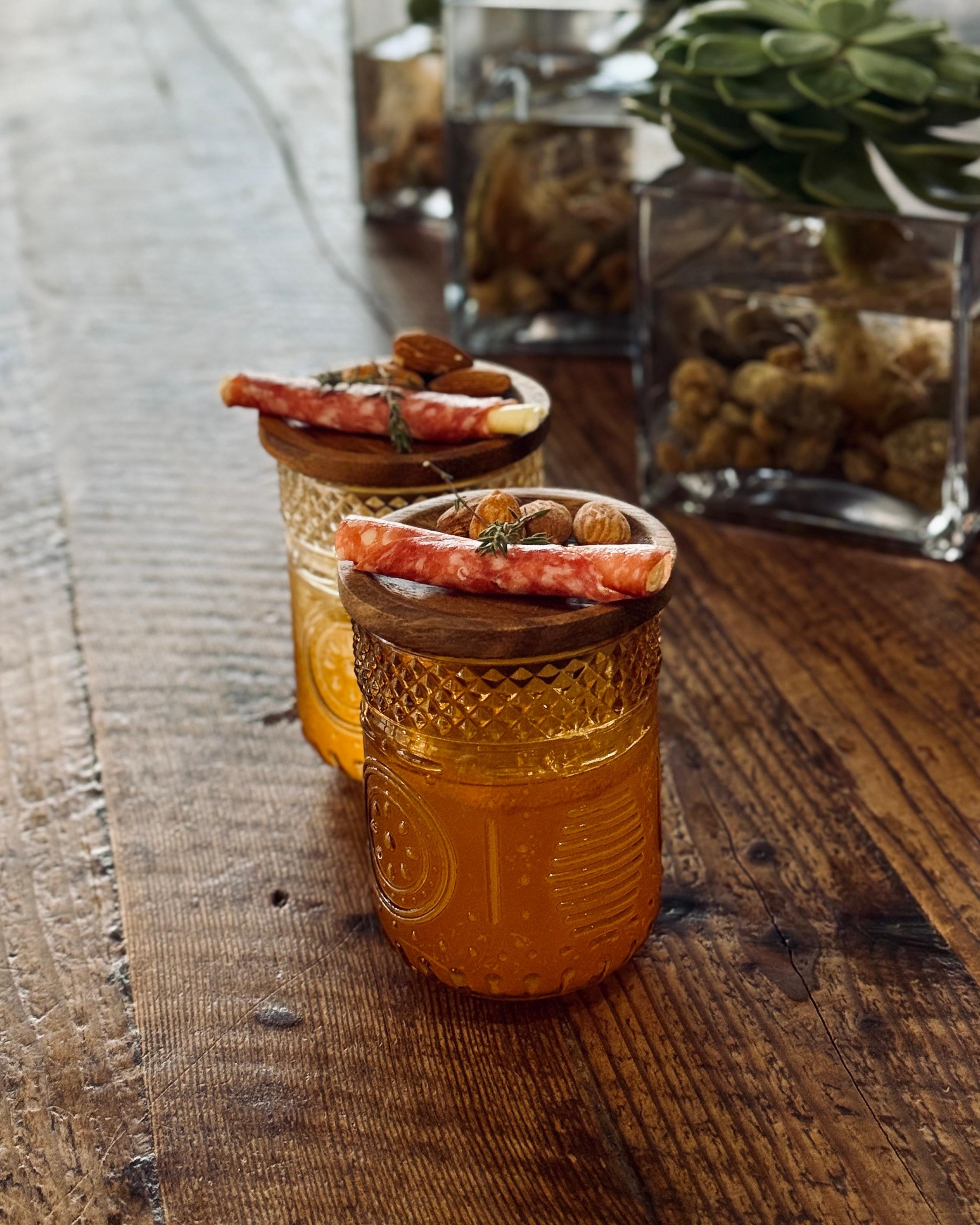 Two small glass jars with decorative lids on a wooden surface with plants in the background.