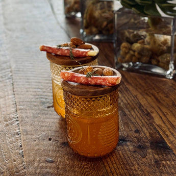 Two small glass jars with decorative lids on a wooden surface with plants in the background.