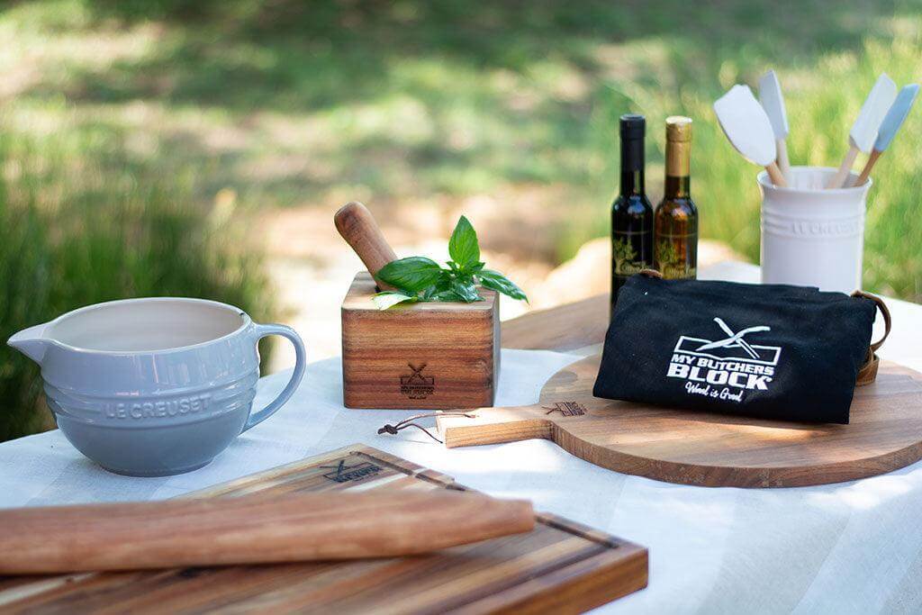 Artisanal kitchen setup including a gray bowl, wooden block, cooking tools, and oils on a rustic table outdoors.