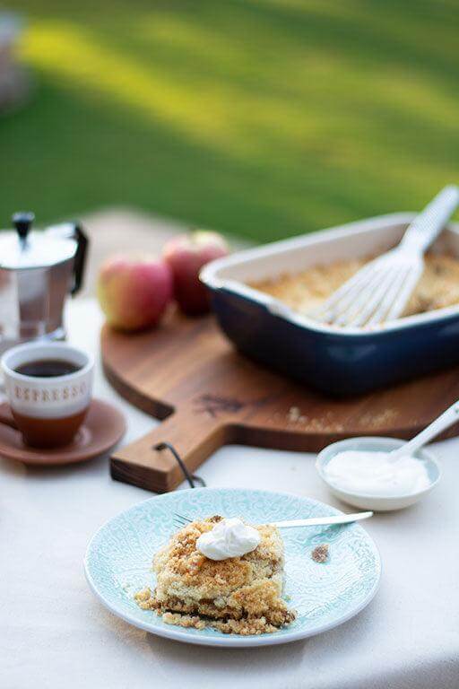 Delicious apple crumble served on a plate with whipped cream, coffee cup, and baking dish in a sunny outdoor setting.