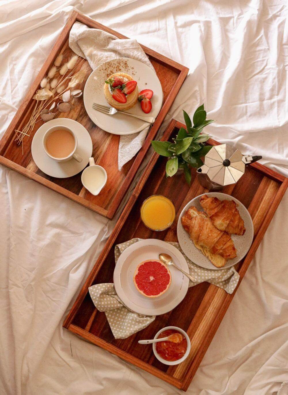 Stylish small serving trays with breakfast items: tea, croissants, fruit, and a coffee pot on a light bedspread.