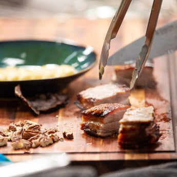 Close-up of sliced barbecue meat on a wooden cutting board with tongs and a bowl of sides.