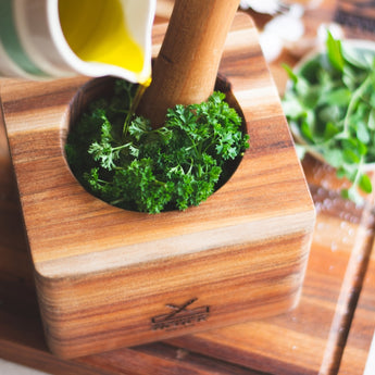 Fresh green parsley being crushed in a wooden mortar with olive oil poured from a jug.