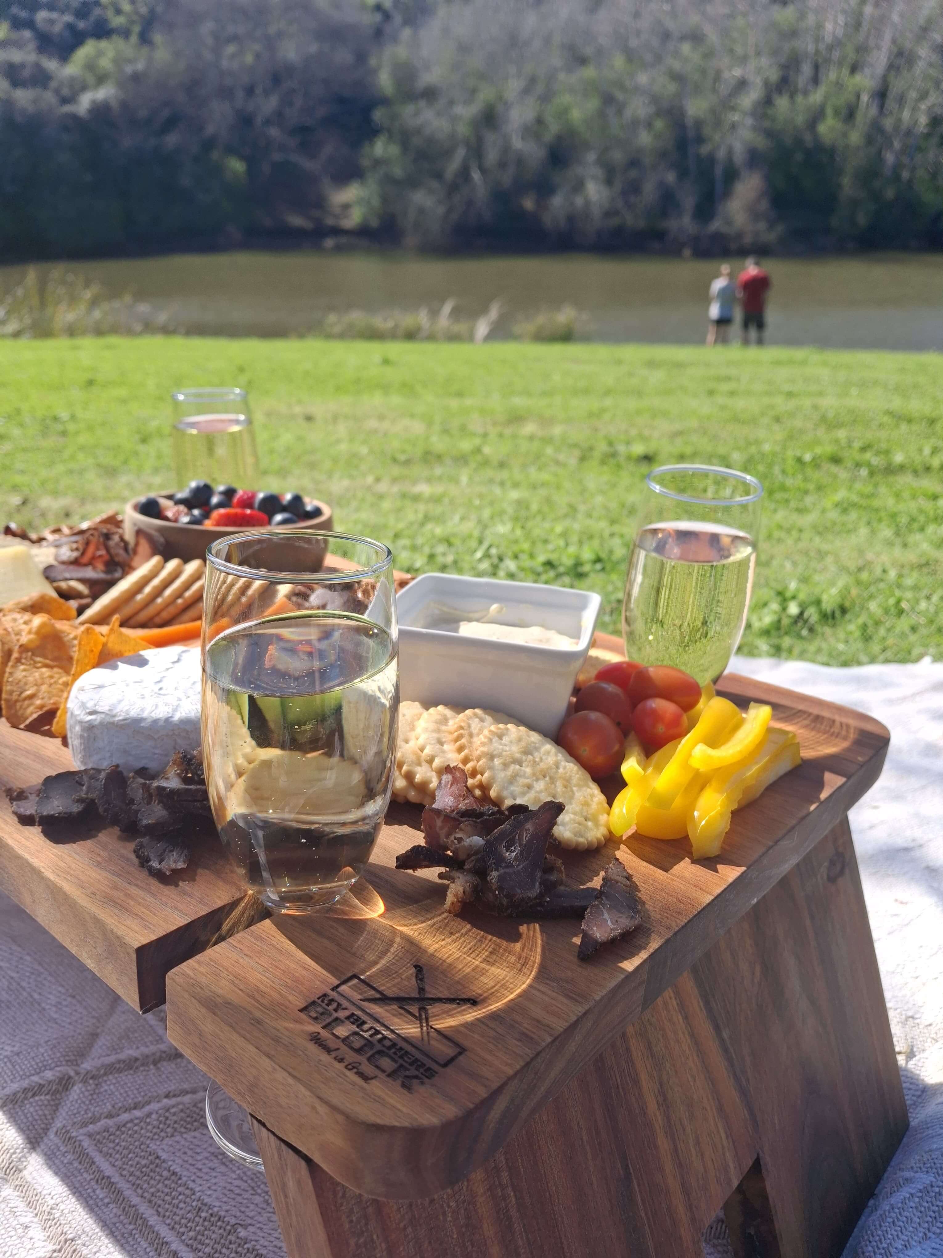 My Butchers Block Grazing Table featuring cheese, fruits, and drinks by a river, perfect for stylish outdoor gatherings.