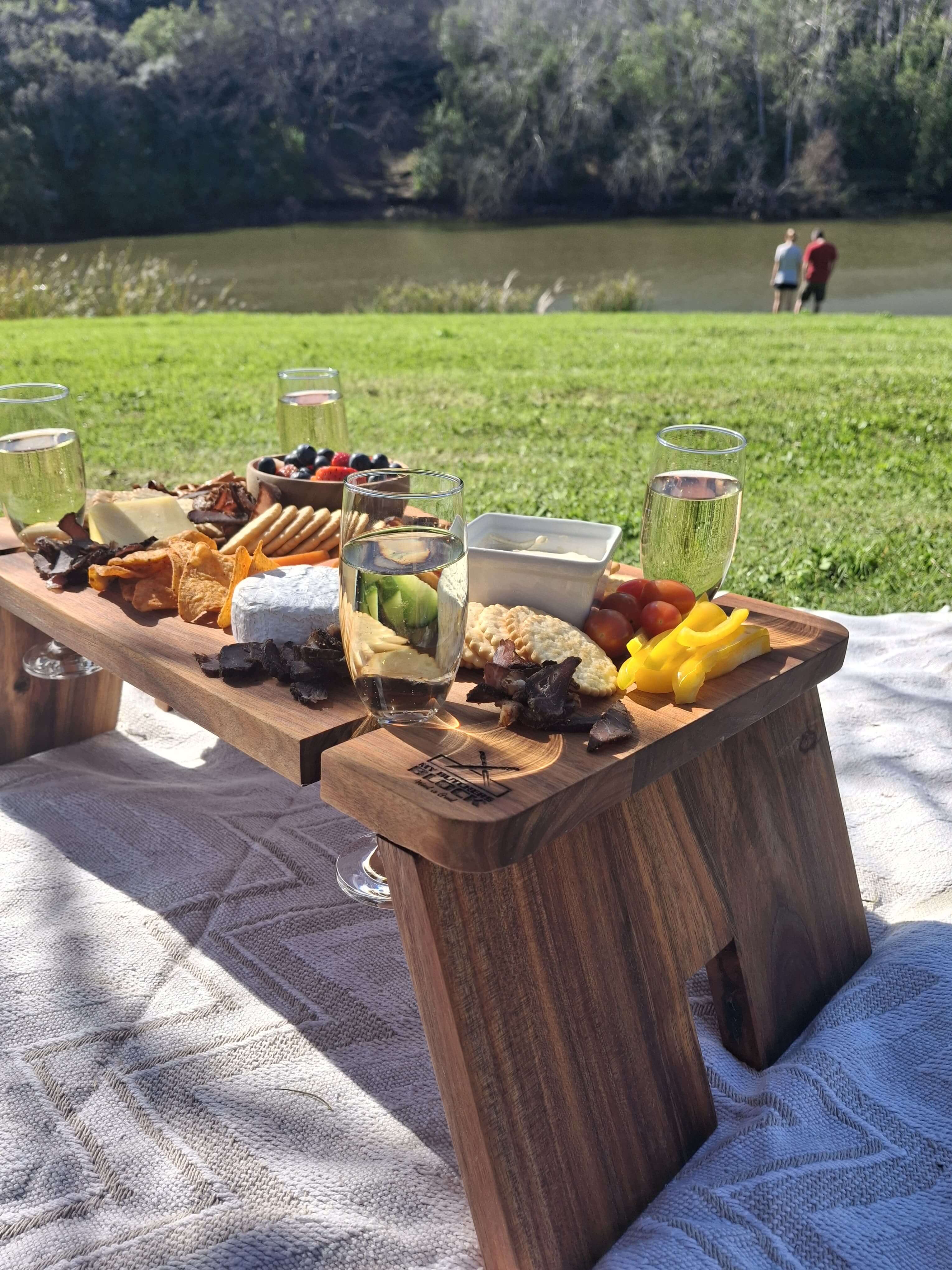 My Butchers Block Grazing Table set outdoors by a scenic lake, featuring a variety of gourmet cheeses, fruits, and drinks.