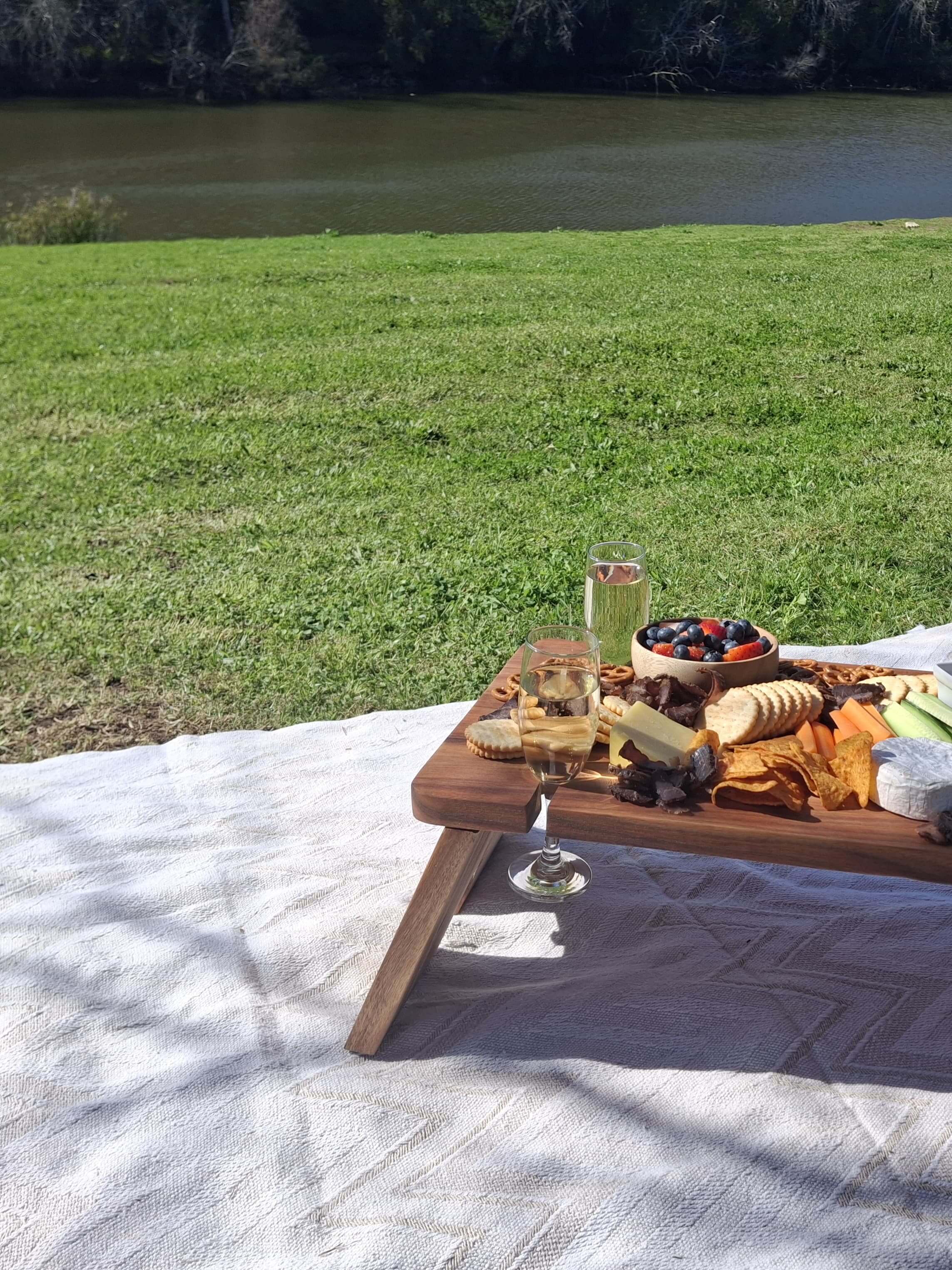 Stylish grazing table with assorted cheese, bread, and wine on a picnic blanket by the riverbank.