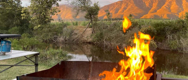 A braai setup with flames in a metal grill surrounded by nature and mountains at sunset.