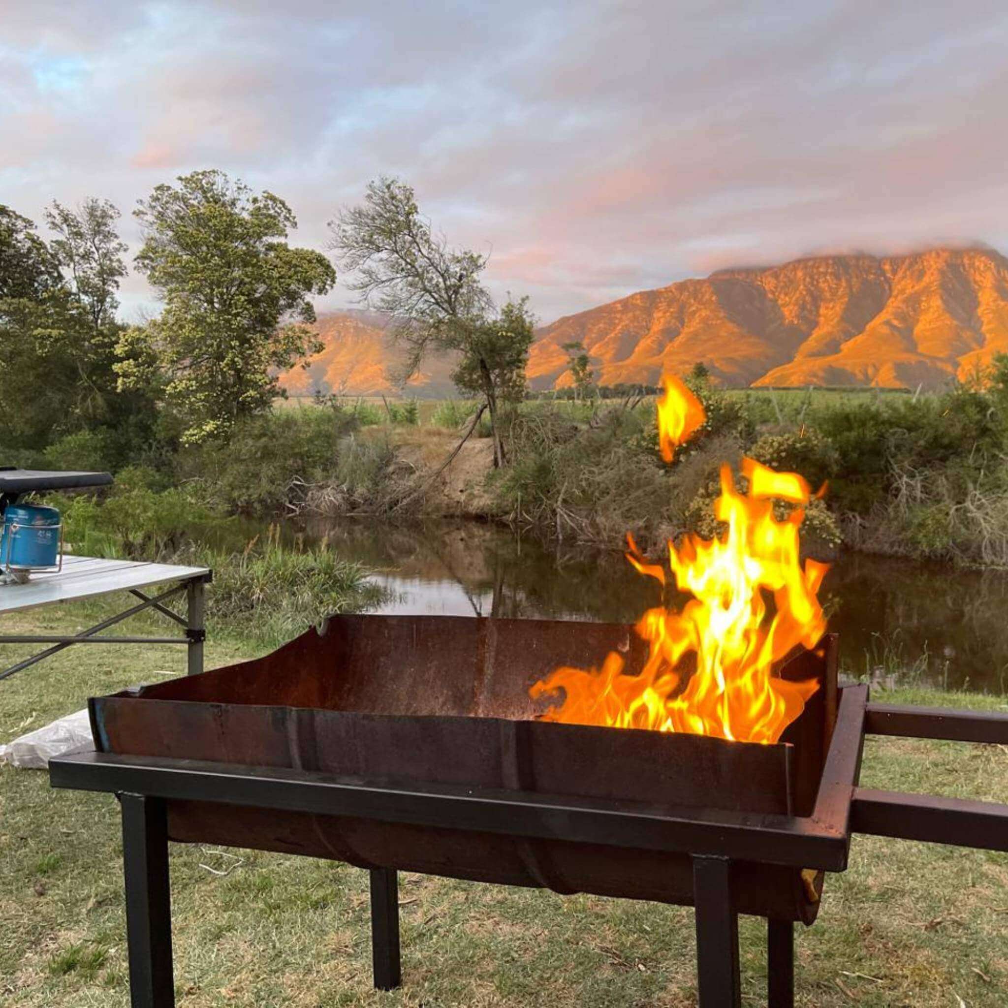 A braai setup with flames in a metal grill surrounded by nature and mountains at sunset.