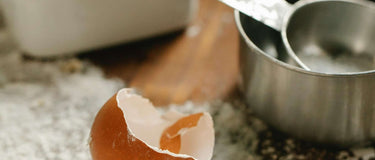Close-up of broken eggshells, measuring cups, and a bowl on a floured kitchen countertop.