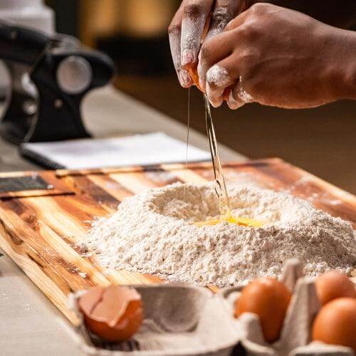 Cracking an egg into a pile of flour on a wooden chopping board in a kitchen setting.