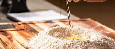 Cracking an egg into a pile of flour on a wooden chopping board in a kitchen setting.