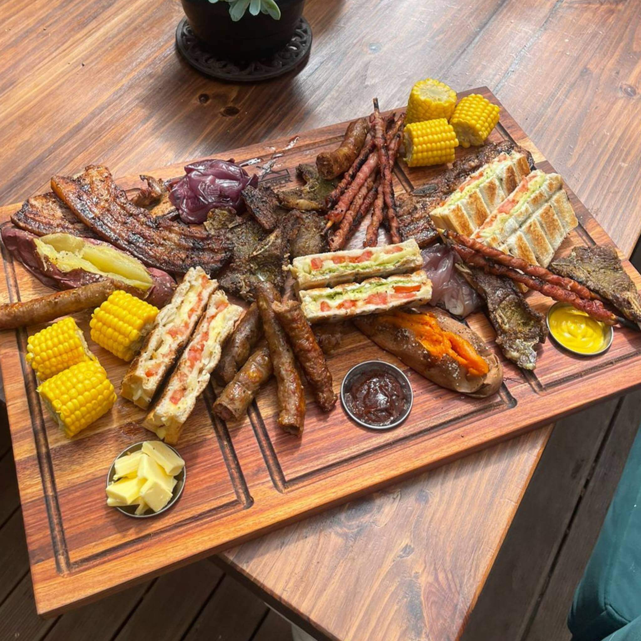 A beautifully arranged braai platter featuring meats, grilled corn, and toasted sandwiches on a wooden board.
