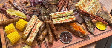 A beautifully arranged braai platter featuring meats, grilled corn, and toasted sandwiches on a wooden board.