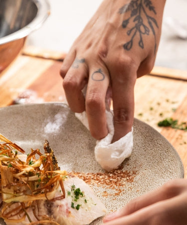 Chef plating a gourmet dish with herbs and spices on a white plate, showcasing culinary skills and creativity.