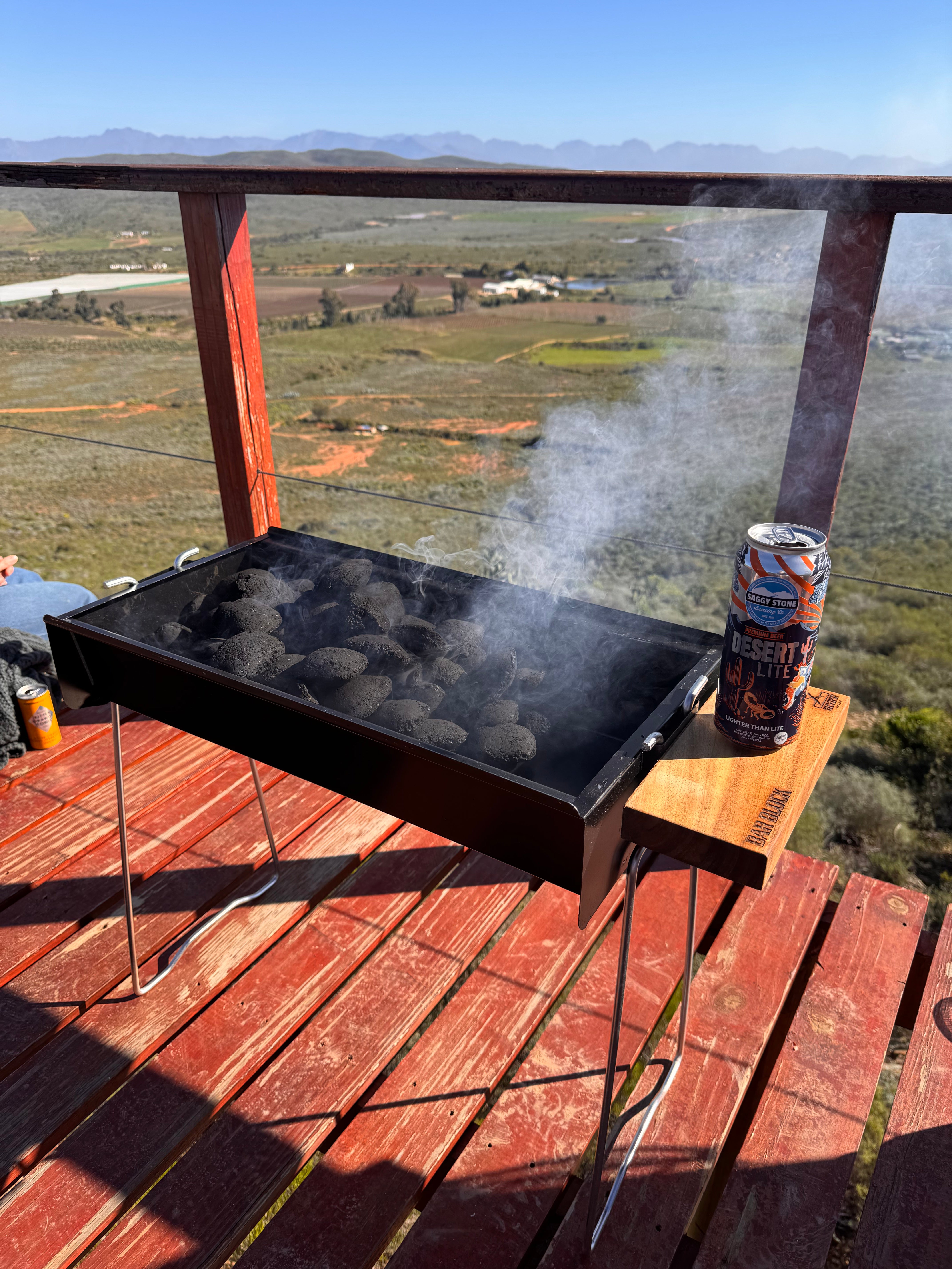 Grill with hot coals and a can of beer on a wooden deck overlooking a scenic landscape.