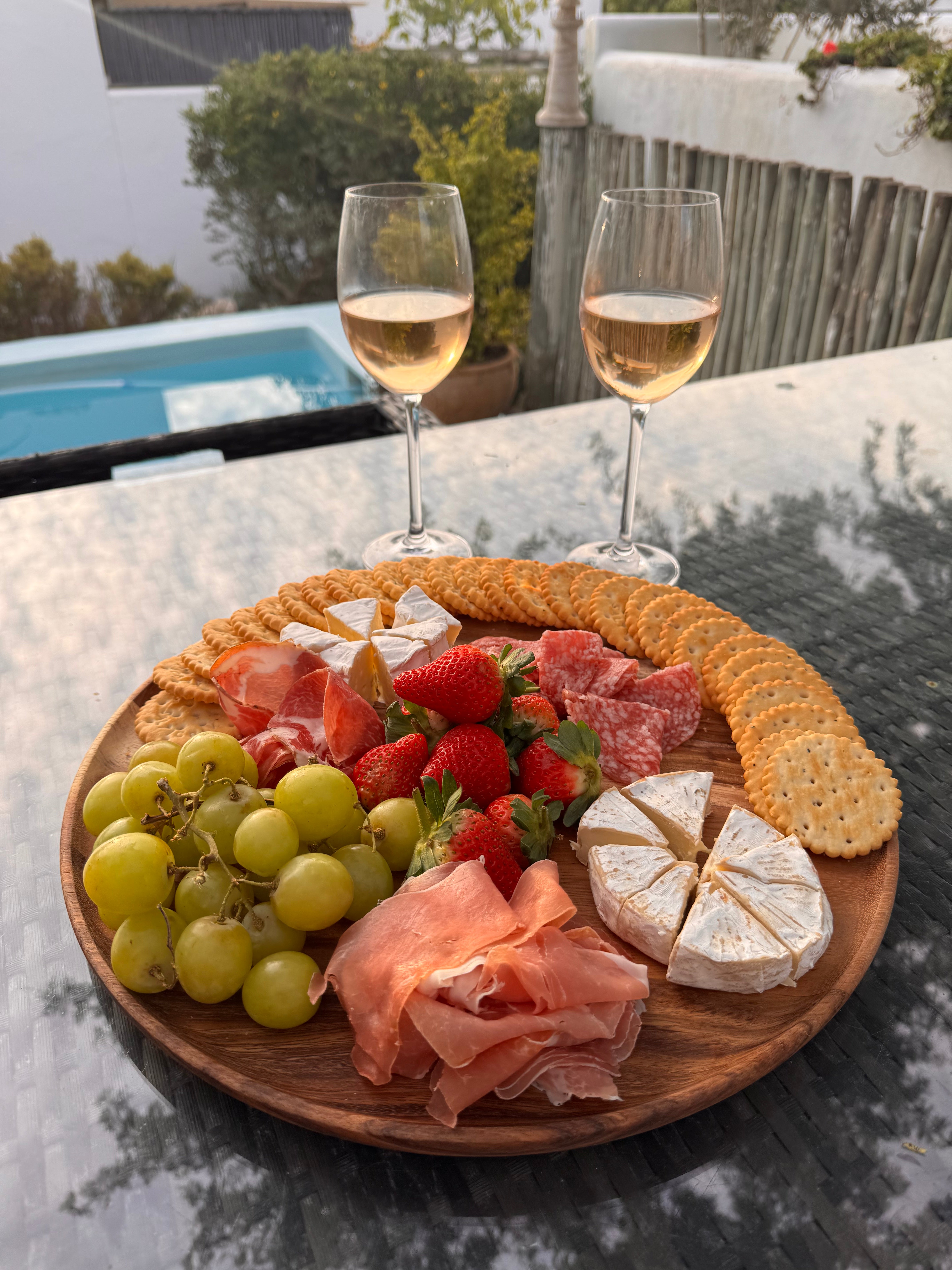 Wooden platter with assorted meats, cheeses, fruits, and crackers on a patio table.