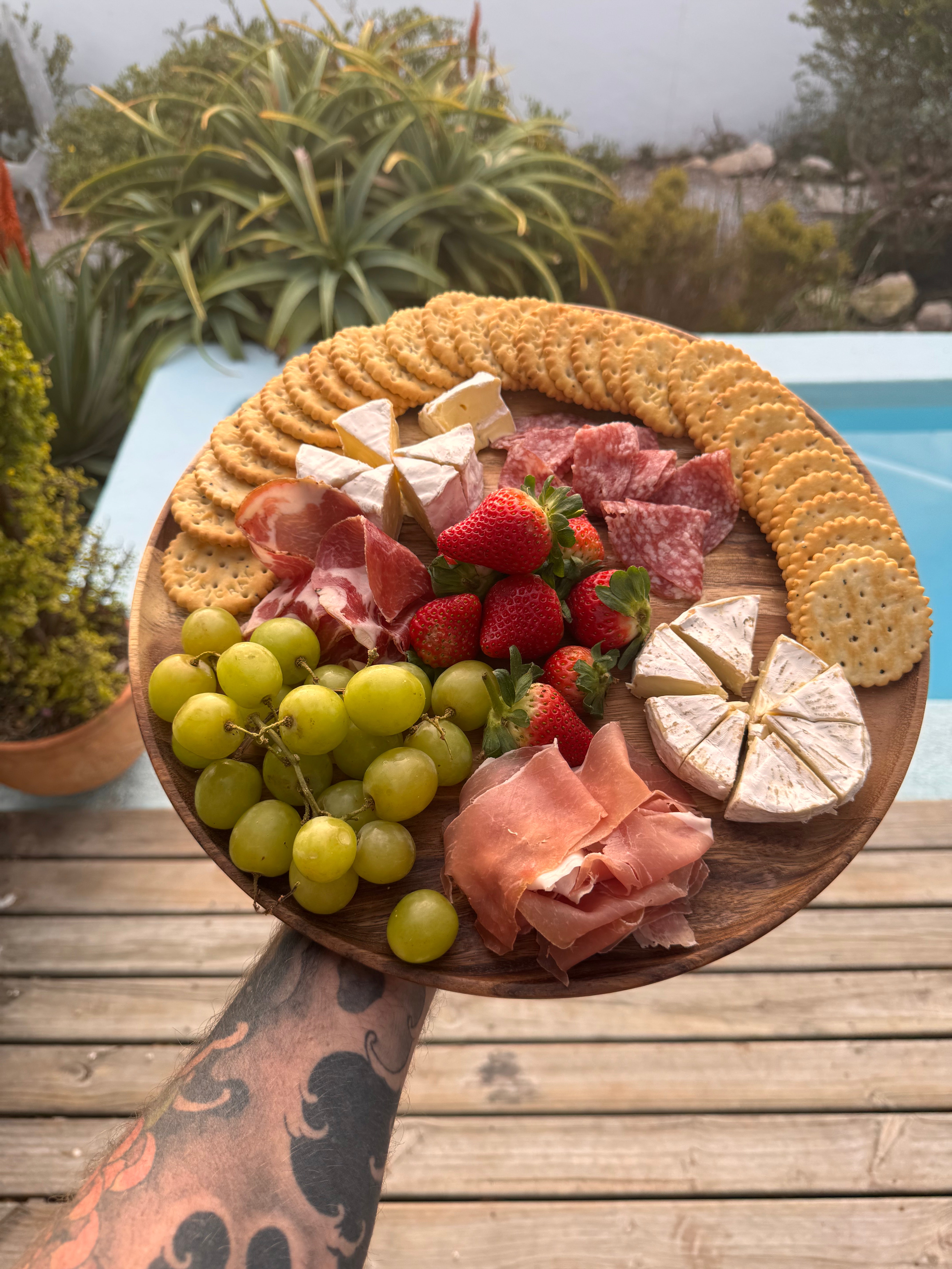 Wooden platter with assorted meats, cheeses, fruits, and crackers on a wooden table outdoors.