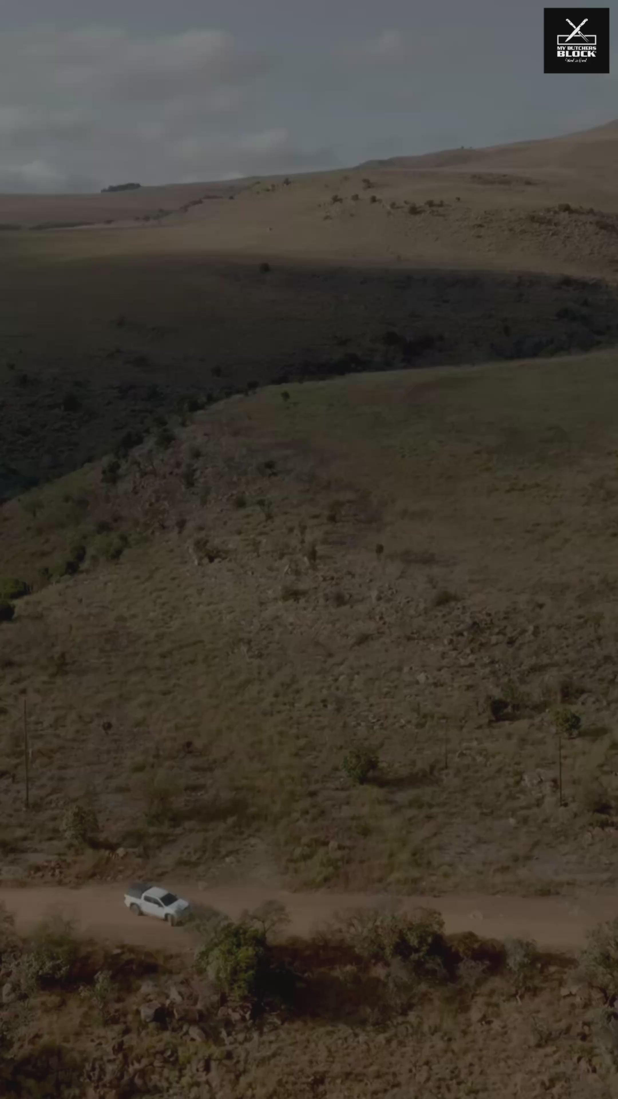 Aerial view of a white car on a dirt road surrounded by green hills and sparse vegetation.